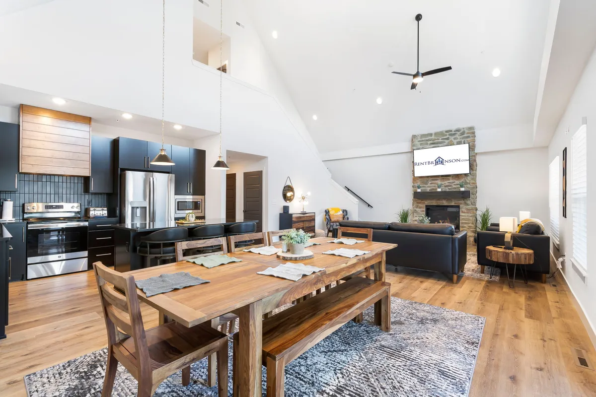 A bright, open-concept space with a modern kitchen, sizable dining table, and inviting living area with a stone fireplace. Large windows bring in light and provide glimpses of the outdoors, reinforcing the open, communal feel.
