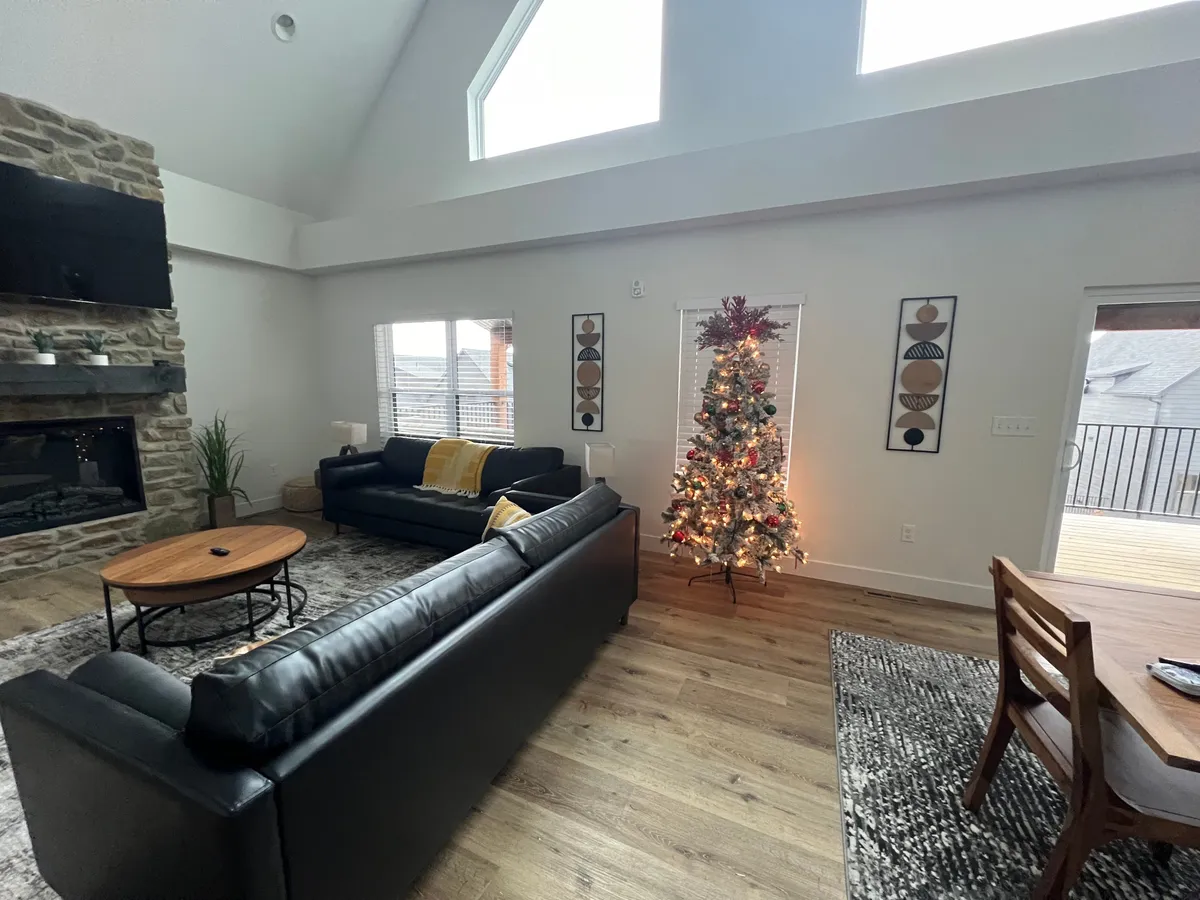 A warmly decorated interior living space featuring a glowing fireplace, a beautifully adorned Christmas tree, a comfortable leather sofa, and a dining area. Sunlight streams through the windows, infusing the room with a festive yet cozy ambiance ideal for winter gatherings and holiday celebrations.