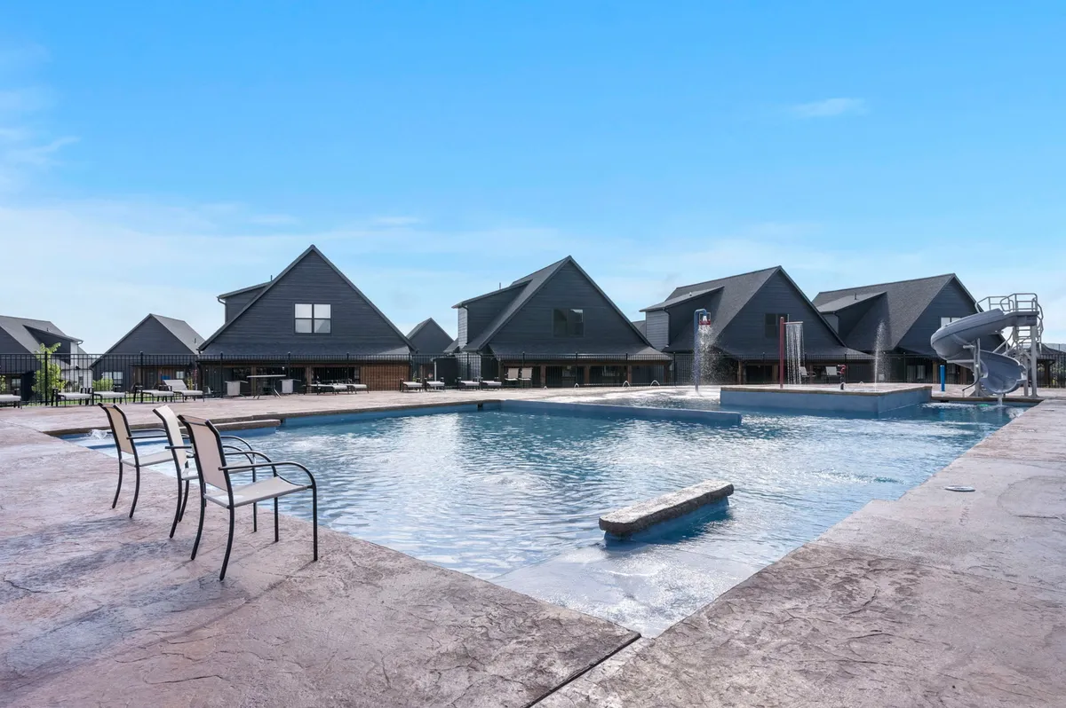 An exterior shot of a community swimming pool, complete with a water slide, surrounding buildings, and plenty of lounge chairs. Children are playing in the water, creating a sense of fun and family-friendly relaxation.