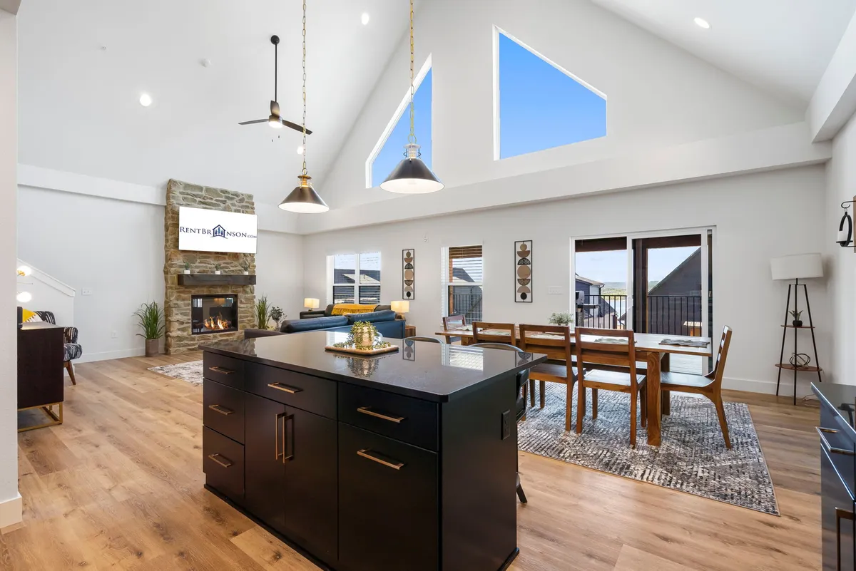 A spacious open-plan main living area featuring a large stone fireplace as the focal point, with plush seating arranged for conversation and relaxation. The space seamlessly connects to a generous dining table and a modern kitchen island, all illuminated by natural light.