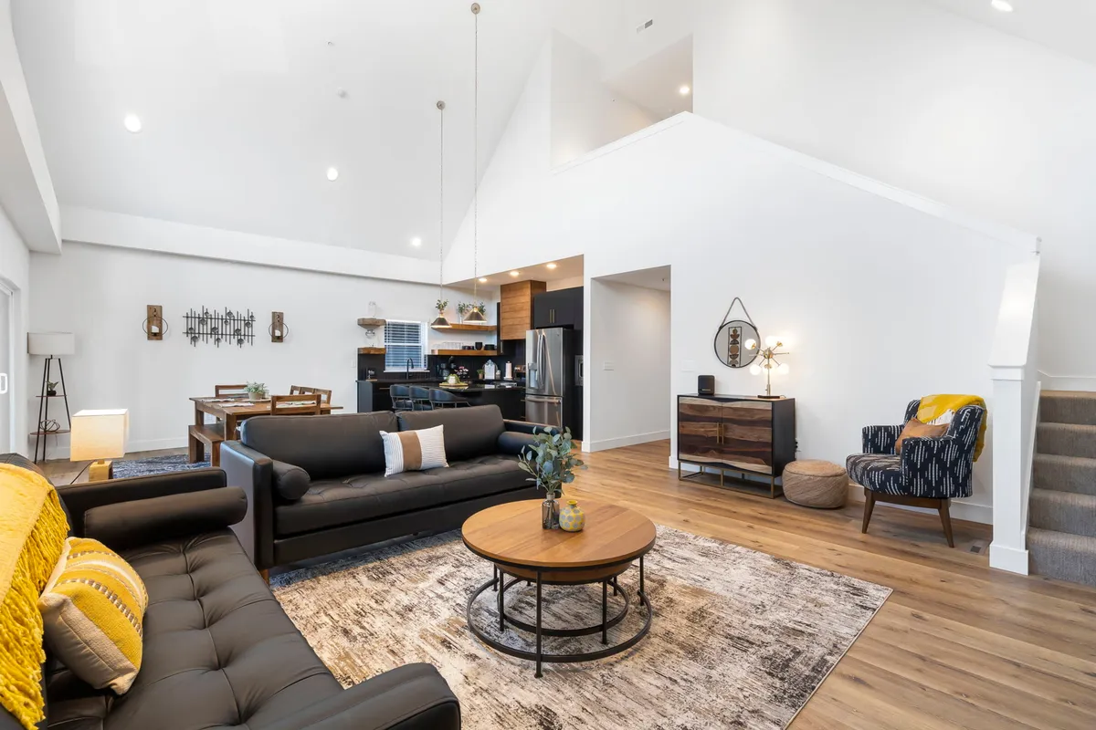 A view from another angle of the social hub, displaying comfortable sofas, a central coffee table, a dining area, a modern kitchen, and a staircase leading upstairs. The layout is open and spacious, designed for both relaxation and entertaining.